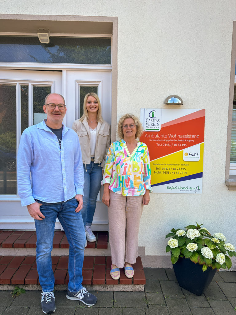 Drei Personen stehen vor einem Gebäude neben einem Schild mit der Aufschrift "Caritas Verein Altenoythe e.V. Ambulante Wohnassistenz"; im Vordergrund ein Mann in blauer Jeans und hellblauem Hemd, dahinter eine junge Frau mit blonden Haaren, beige Jacke und Jeans sowie eine Frau mit lockigen blonden Haaren, bunter Bluse und beiger Hose; rechts ein großer Topf mit weißen Hortensien.
