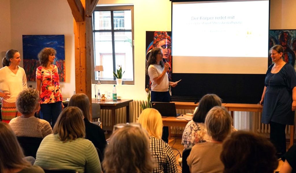 Frau hält Rede mit Mikrofon vor Leinwand in kleinem Seminarraum, Publikum sitzt auf Stühlen und hört zu.